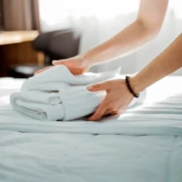 Detailaufnahme einer Hand, die frische, weiße Handtücher auf einem Bett in einem hellen Raum faltet, ein Beispiel für die Sorgfalt und Aufmerksamkeit, die S.K. Reinigungsservice Leipzig e.K. in ihre Arbeit einbringt.